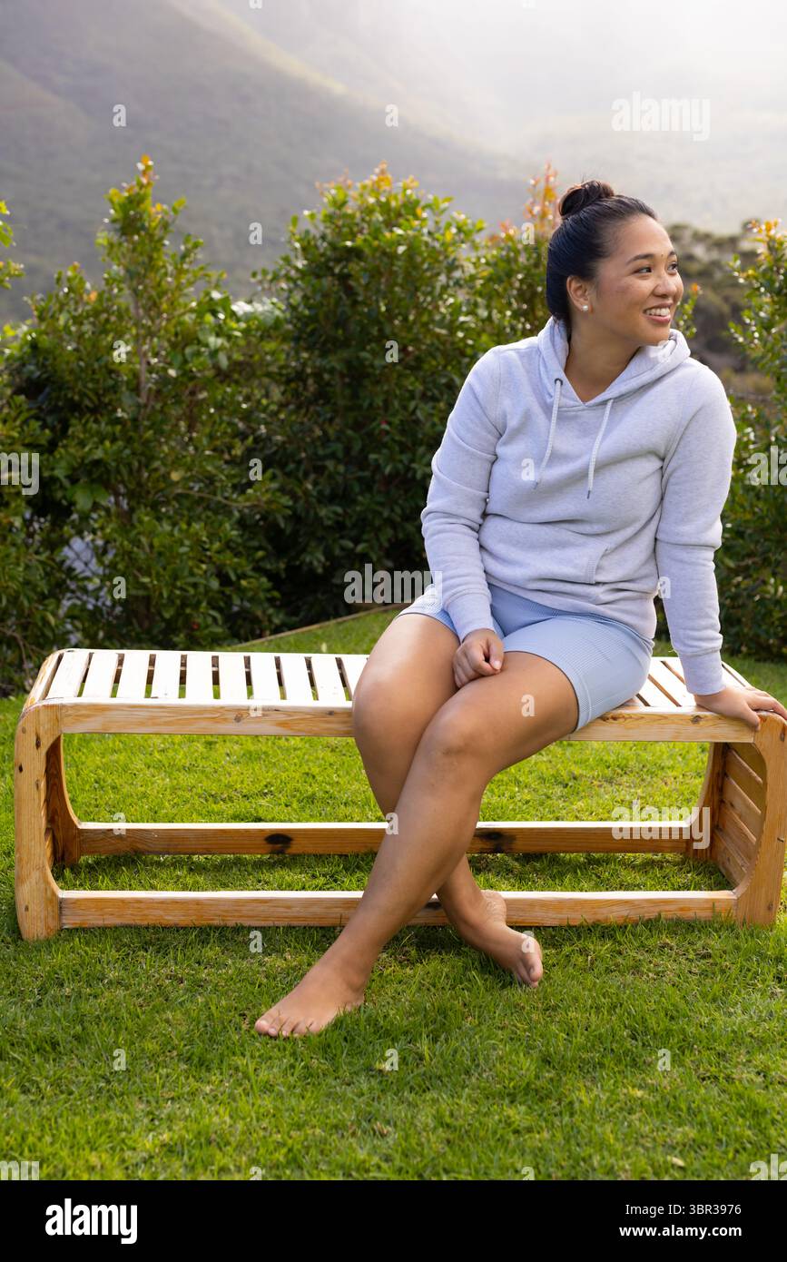 Donna asiatica seduta a piedi nudi su una panchina di legno a doghe su prato erboso con siepe rifilata, copia spazio Foto Stock