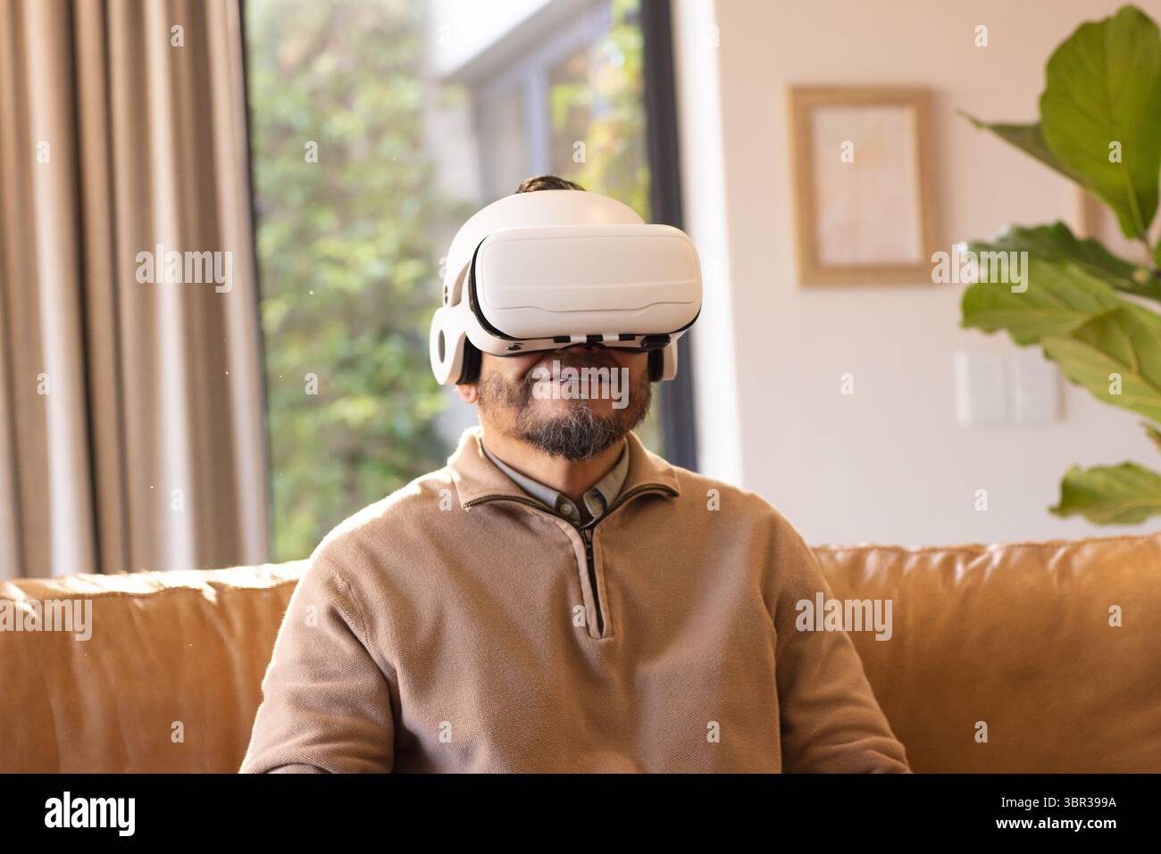 Visore per realtà virtuale bianco seduto su un divano in pelle abbronzata vicino a una finestra illuminata dal sole che mostra il fogliame verde Foto Stock