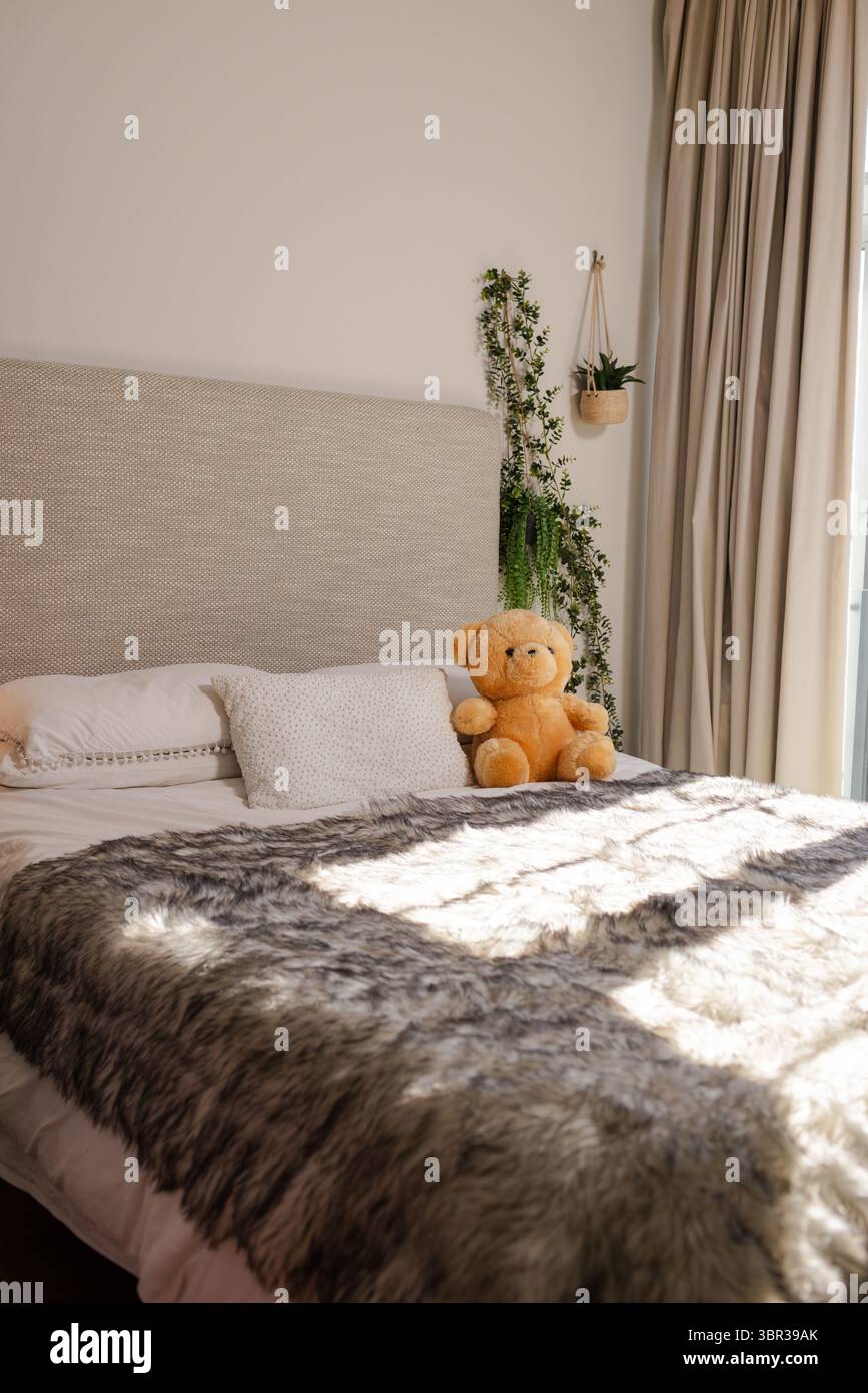 Letto queen size con getti in pelliccia sintetica e orsacchiotto riposante, luce del sole che scorre attraverso le tende Foto Stock