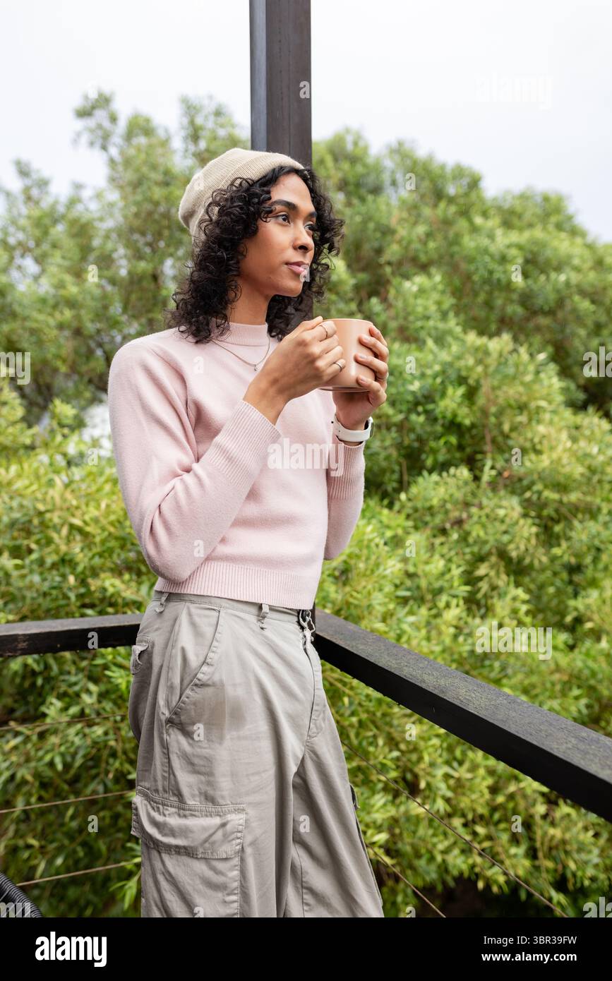 Persona non binaria che indossa il berretto, tiene in mano una tazza di ceramica e osserva le ringhiere del balcone, spazio per fotocopie Foto Stock