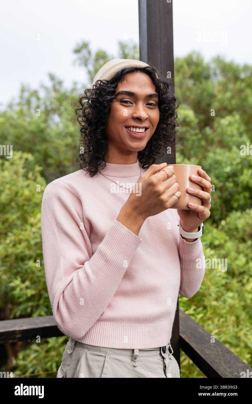 Persona non binaria in piedi sul ponte con maglione rosa chiaro, tenendo la tazza marrone vicino agli arbusti verdi Foto Stock