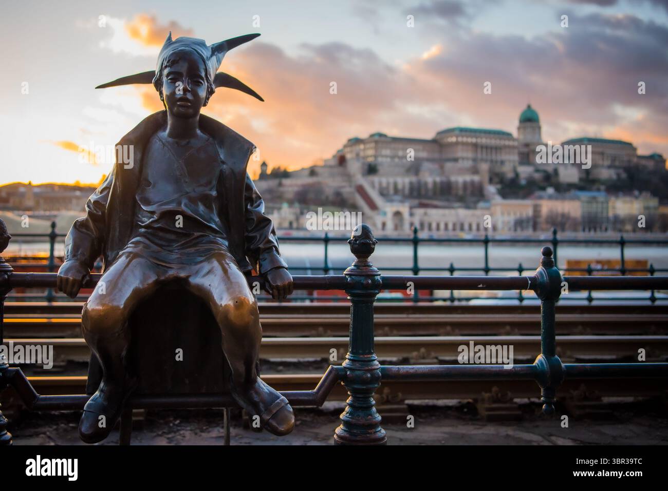 Vista della statua in bronzo di un ragazzo di Pest con orecchie a punta e un cappotto, sullo sfondo del castello di Buda al tramonto, Budapest, Ungheria. Foto Stock