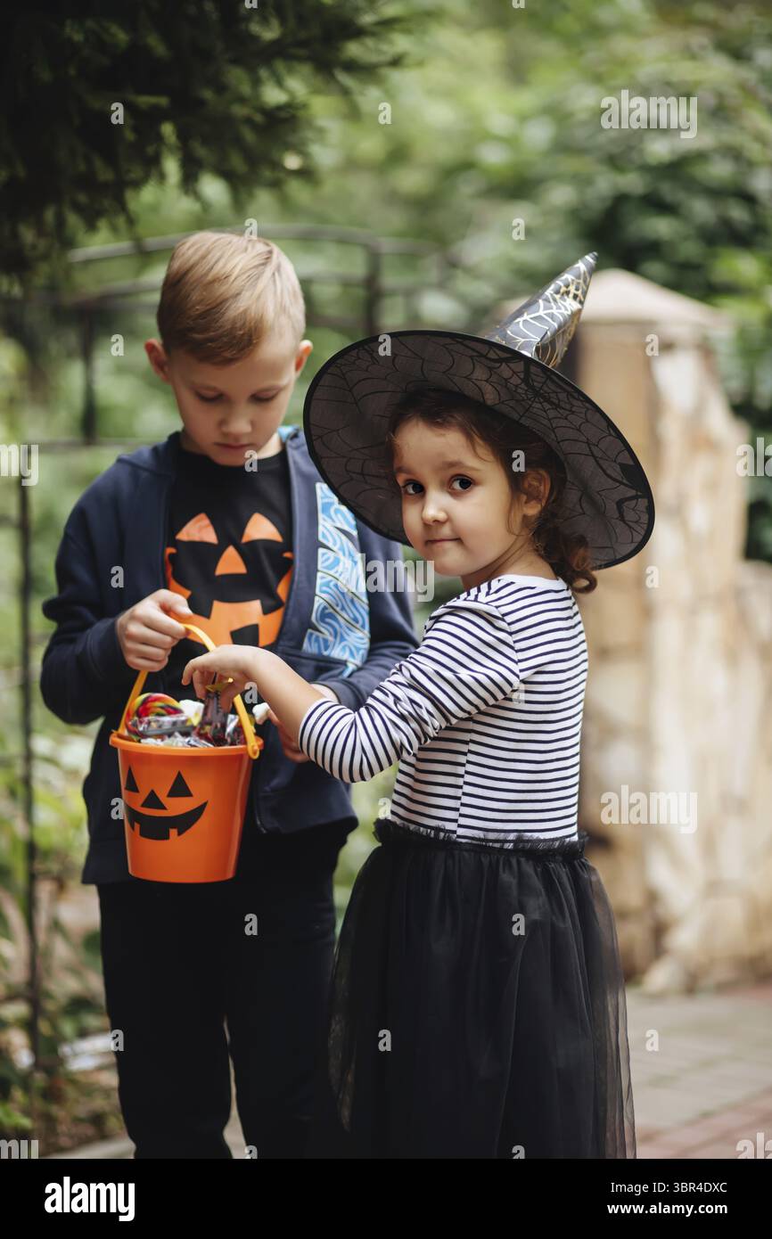 Kleine niedliche Maedchen in Hexenkostuem und Junge haelt Jack-o-Lantern Kuerbis Eimer mit Suessigkeiten und Suessigkeiten und schmeckt Suessigkeiten. Foto Stock