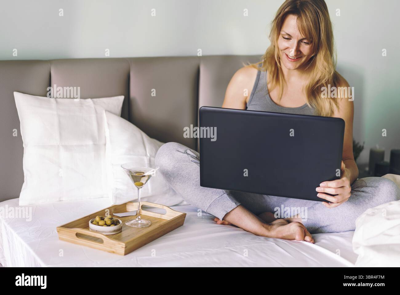 Apprendimento a distanza formazione online o lavoro o chiacchierare con gli amici. Donna felice sdraiata in un letto, che usa un laptop e beve martini Foto Stock