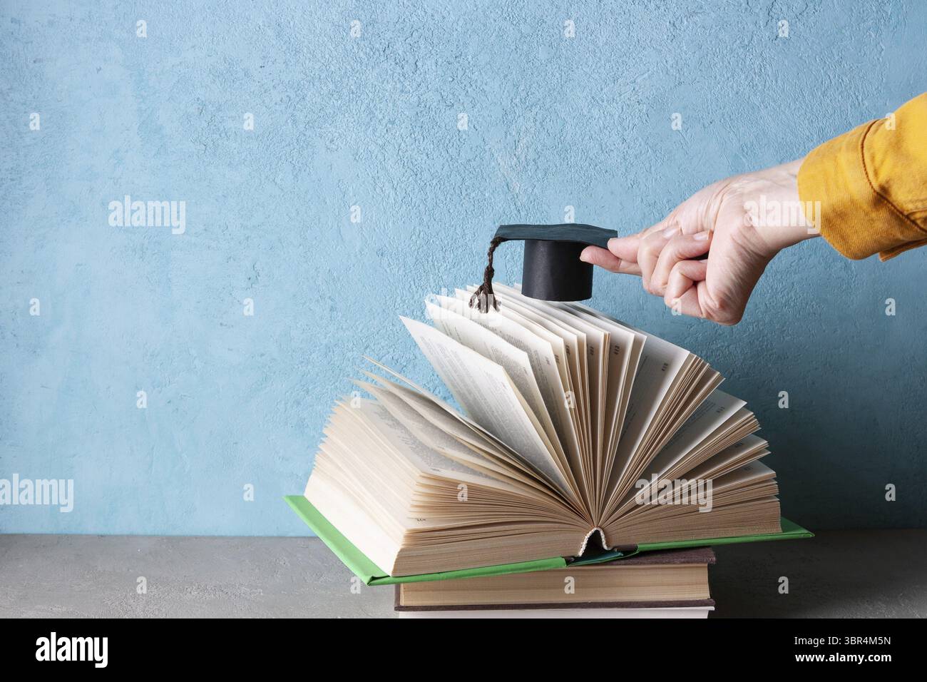 La mano di una donna che tiene un cappello da studente di carta sopra un libro aperto Foto Stock