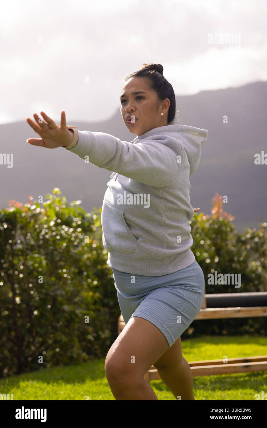 Donna asiatica che indossa una felpa con cappuccio grigia, pantaloncini blu che tengono in posa il guerriero yoga su prato per panchina, siepe Foto Stock