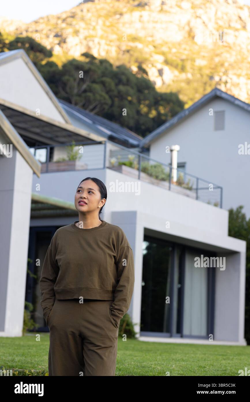 Donna asiatica in piedi su un prato verde di fronte a una moderna casa a due piani con piantatrici di balconi Foto Stock