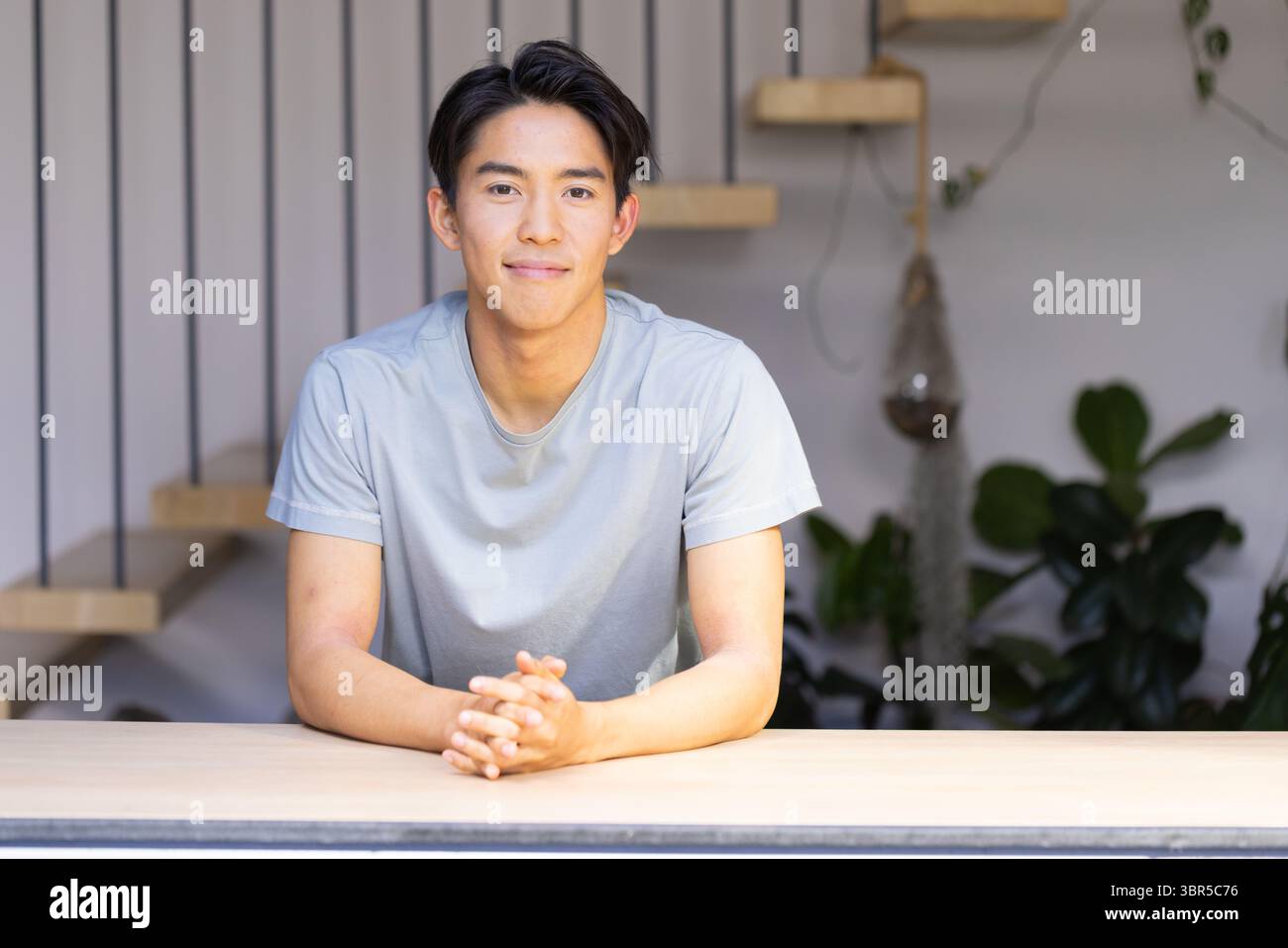 Seduto a un tavolo di legno chiaro in interni minimalisti, uomo asiatico che stringe le mani vicino alla scala Foto Stock
