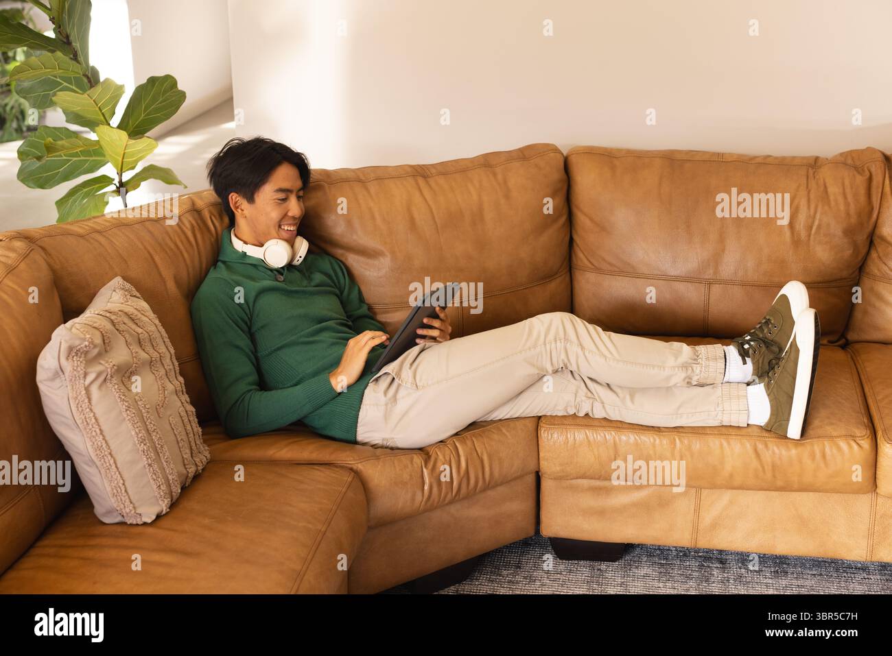 Uomo adulto asiatico reclinato su un divano in pelle abbronzata nell'angolo del soggiorno, utilizzando un tablet con cuffie Foto Stock