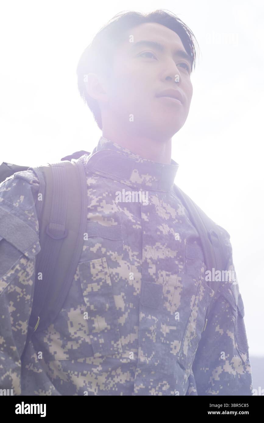 Uomo asiatico in piedi, che guarda verso l'alto sotto il sole in uniforme mimetica digitale con zaino tattico Foto Stock