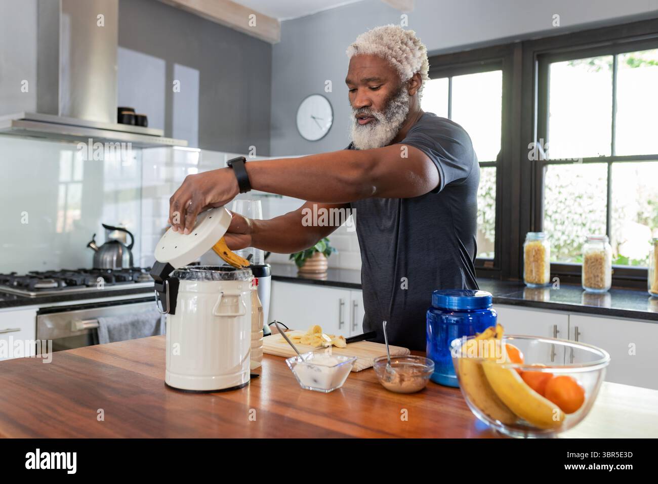 Un anziano afroamericano che colloca la banana sbucciata nel mixer dell'isola della cucina con polvere proteica Foto Stock