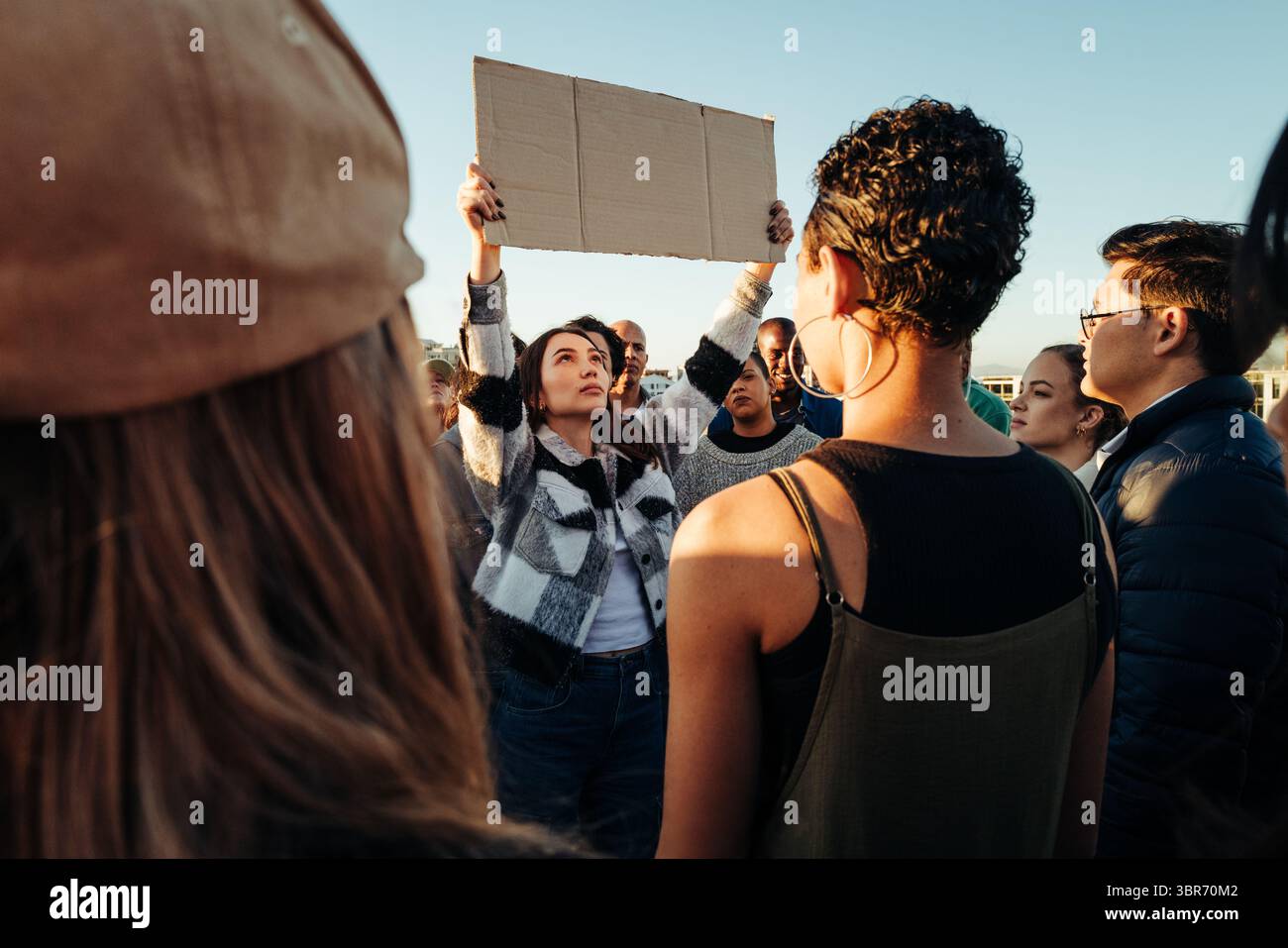 Una giovane donna determinata solleva un cartello di cartone mentre si trova al centro di un raduno di attivisti. La folla è composta da diversi individui che Foto Stock