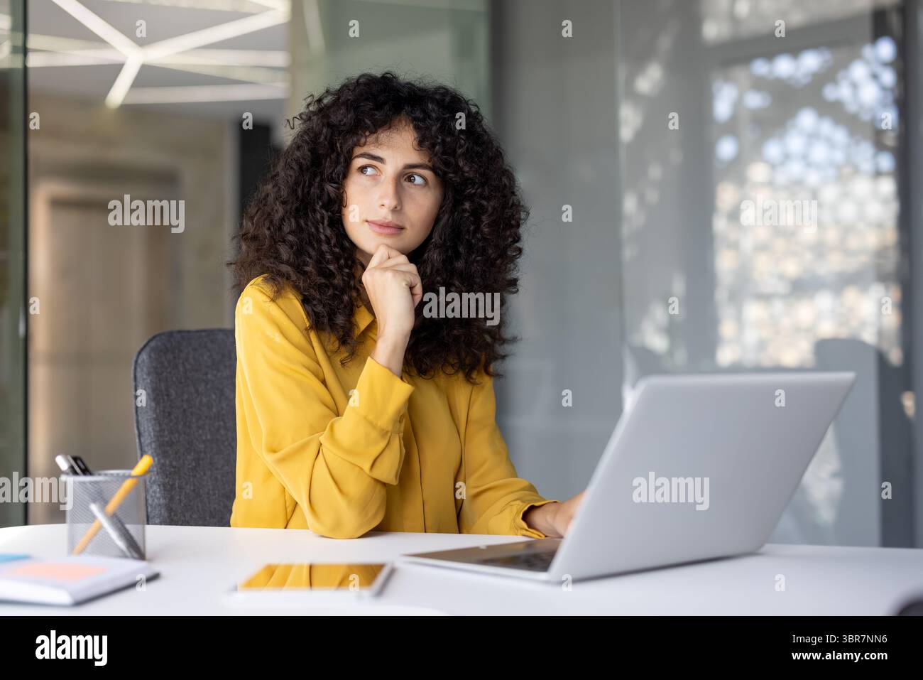 Una donna premurosa con capelli ricci che si stupisce mentre si siede a una scrivania con un computer portatile in un ambiente di ufficio. Foto Stock