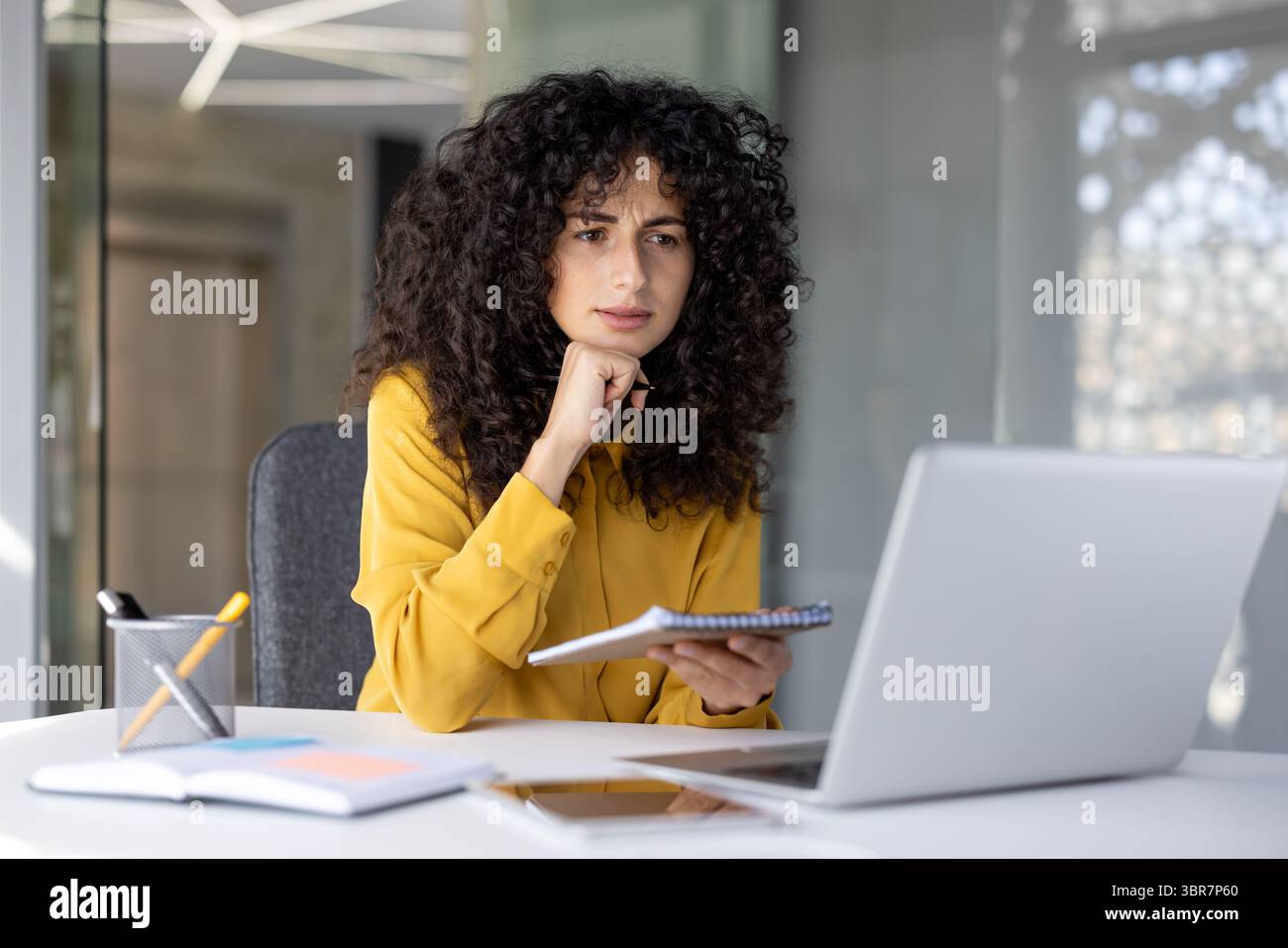 Una donna d'affari premurosa con una camicia gialla, che riflette sulle note e su un notebook in un moderno ambiente da ufficio. Foto Stock