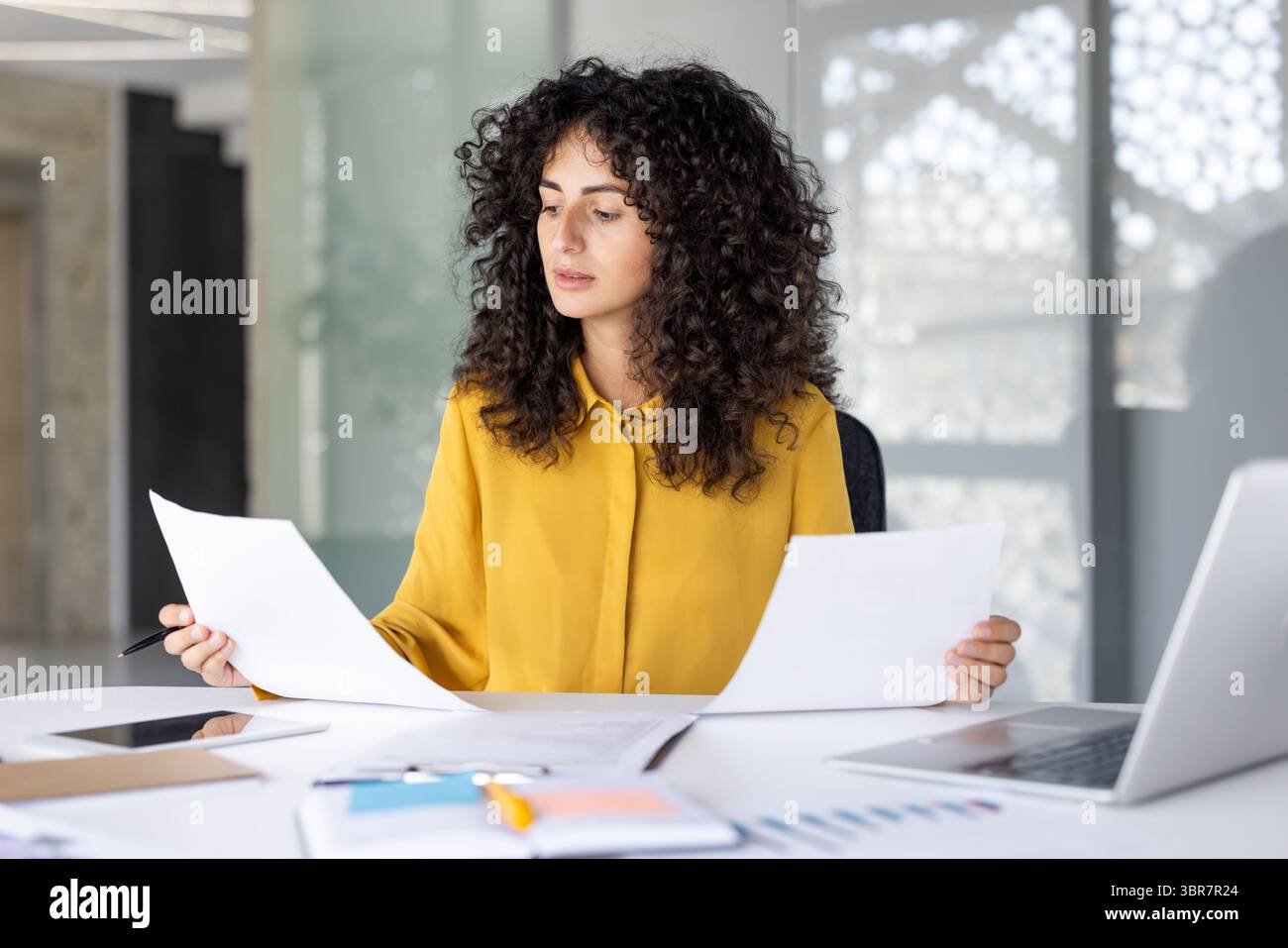 Una donna dai capelli ricci esamina i documenti alla scrivania, mostrandosi concentrata sul suo lavoro in un ambiente luminoso. Foto Stock