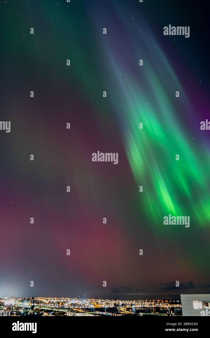Vista dell'aurora boreale danzante in vivaci tonalità verde e viola sopra le scintillanti luci della città, un balletto celeste nel cielo notturno, Reykjavík, Reykjavíkurborg, Islanda. Foto Stock