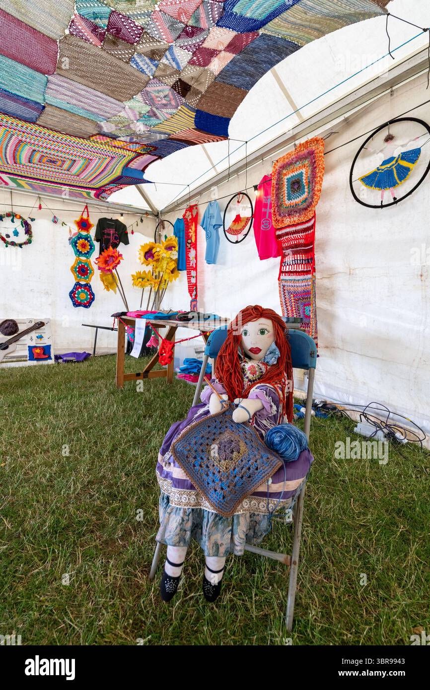Bambola di straccio seduta ad uncinare un tappeto quadrato con una colorata esposizione di capi all'uncinetto e fiori all'interno di una tenda durante un festival musicale Foto Stock