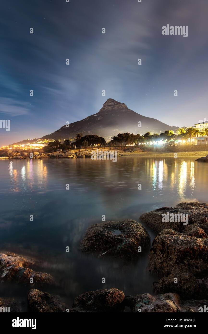 Vista della tranquilla piscina Tidal Pool di Camps Bay che riflette le luci della città sotto la maestosa montagna Lion's Head, con rocce in primo piano che aggiungono profondità, Cape Tow Foto Stock
