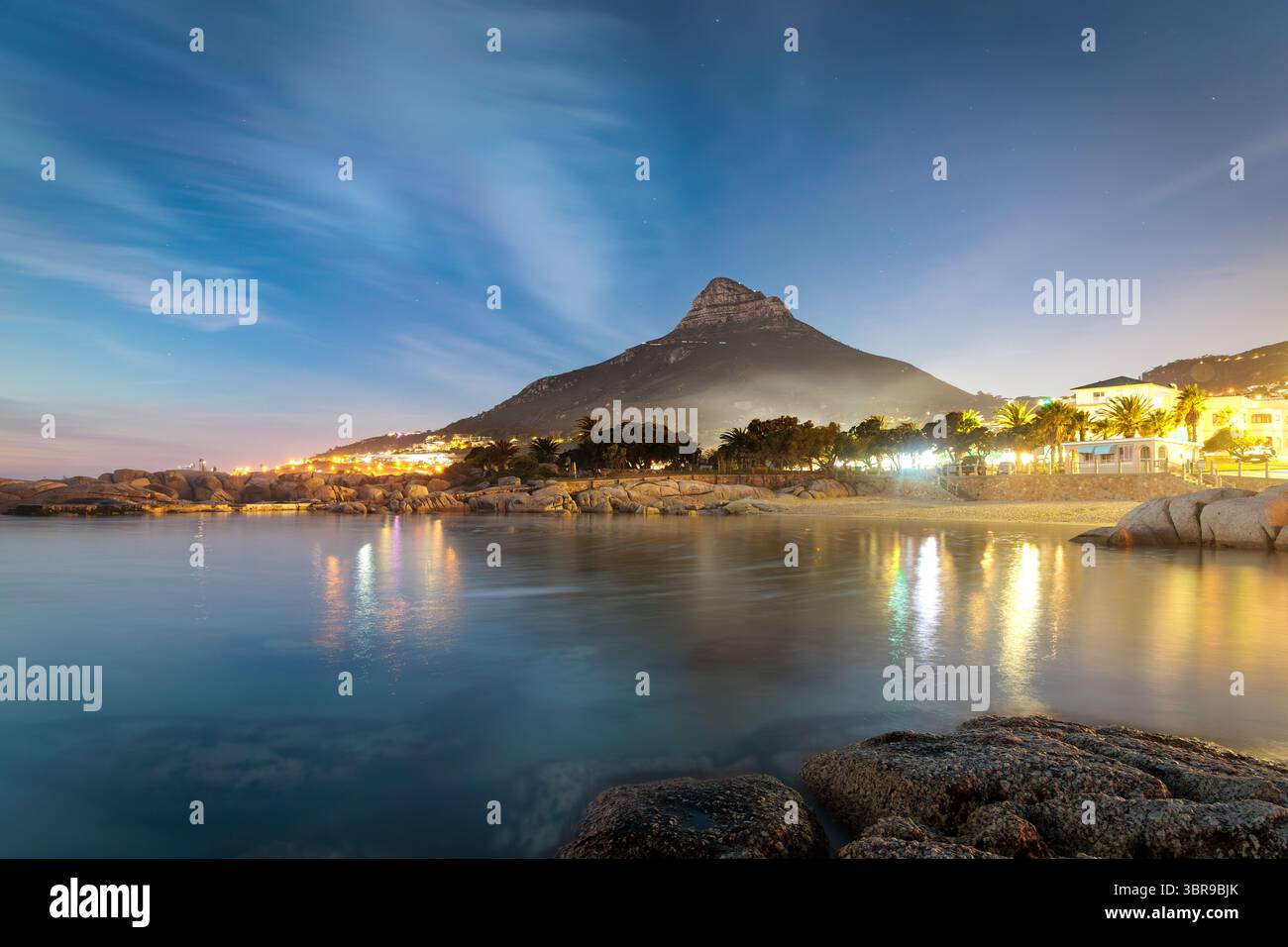 Vista della montagna Lion's Head che proietta una lunga ombra sulla tranquilla baia, con le luci della città che brillano nel tramonto Camps Bay Tidal Pool, città del Capo, Capo Occidentale, Sud Africa. Foto Stock
