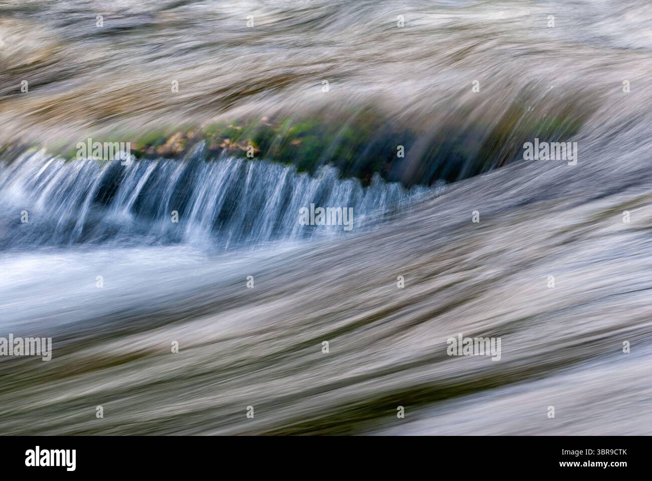 L'acqua limpida cade su pietre lisce, creando ondulazioni e riflessi delicati in un ruscello tranquillo sotto la luce del giorno. Foto Stock