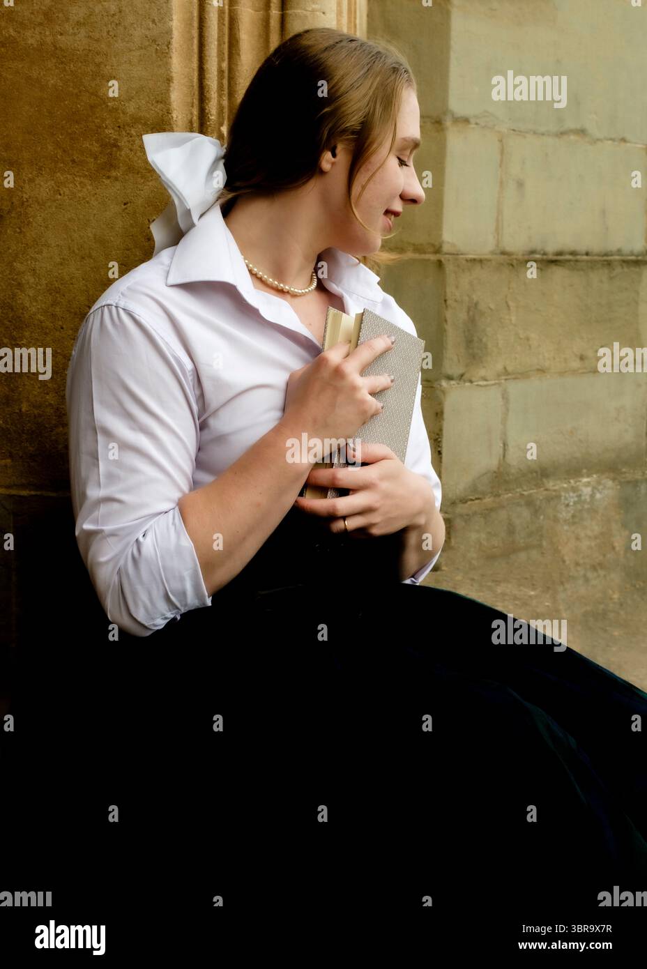 Una giovane donna con una camicia bianca e una collana di perle tiene in mano un libro, appoggiata contro un muro di pietra. Foto Stock