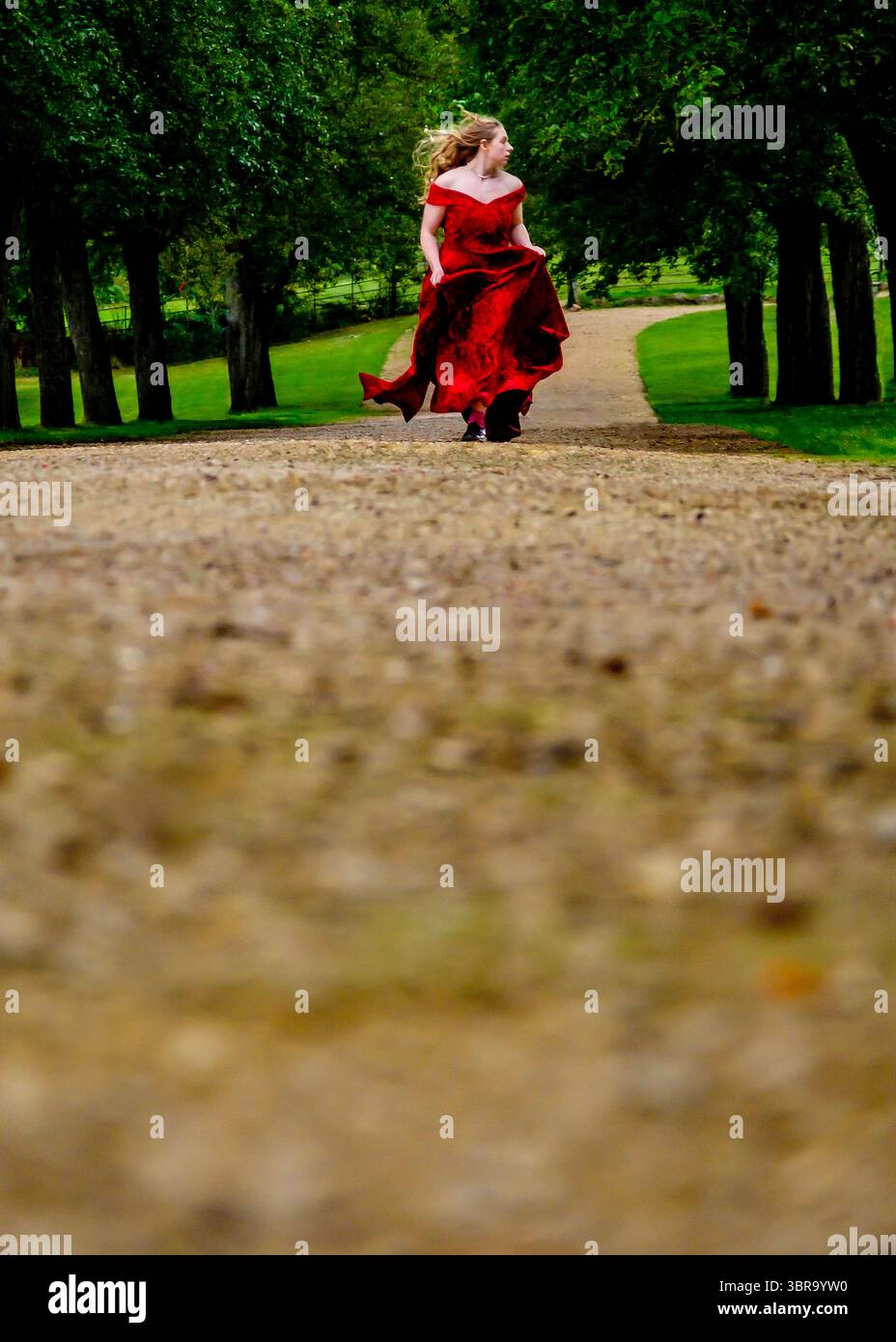 Una donna con un vestito rosso corre lungo un sentiero di ghiaia fiancheggiato da alberi. Foto Stock