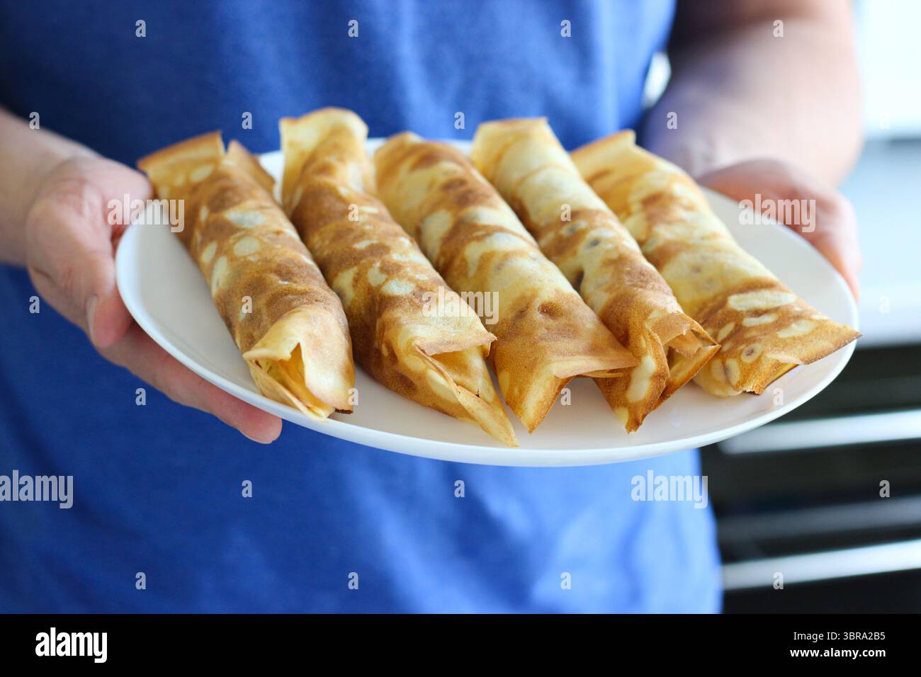 Uomo in camicia blu che regge un piatto bianco con cinque crepe arrotolate ripiene di formaggio. Concentrati sulle mani e sul cibo. Colazione casalinga con atmosfera rustica Foto Stock