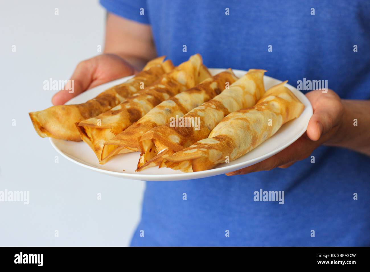 Uomo in camicia blu che regge un piatto bianco con cinque crepe arrotolate ripiene di formaggio. Concentrati sulle mani e sul cibo. Colazione casalinga con atmosfera rustica Foto Stock