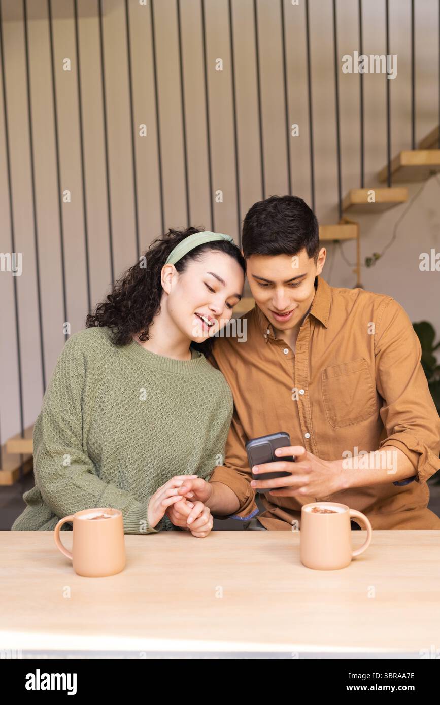 La coppia si siede al tavolo di legno nell'area soggiorno e condivide lo smartphone con tazze da caffè in ceramica Foto Stock