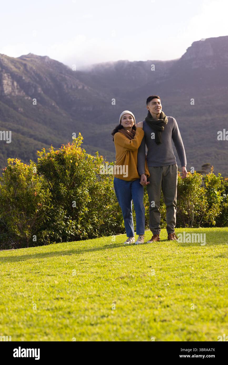 Coppia variegata che tiene le mani nel parco di montagna su un prato verde con berretto e sciarpa in maglia Foto Stock