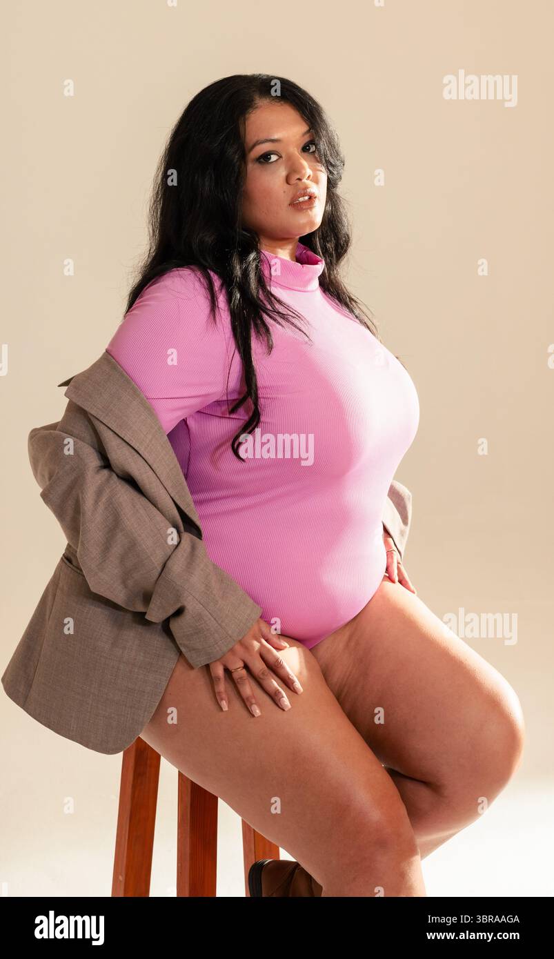 Modello femminile seduto lateralmente su sgabello in legno in studio, con body rosa e blazer grigio Foto Stock