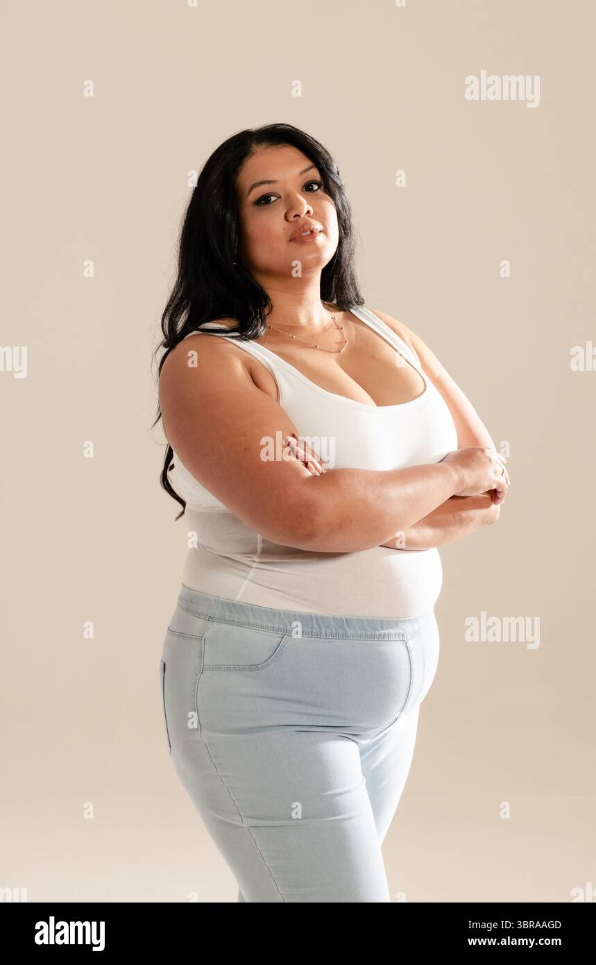 Donna asiatica che posa con le braccia incrociate in studio indossando canotta bianca, jeans blu, gioielli in oro Foto Stock