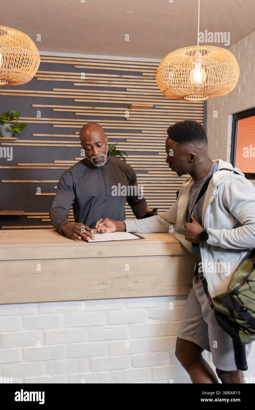 Assistente afroamericano che esamina gli appunti alla scrivania mentre il membro della palestra firma il modulo con la borsa Foto Stock