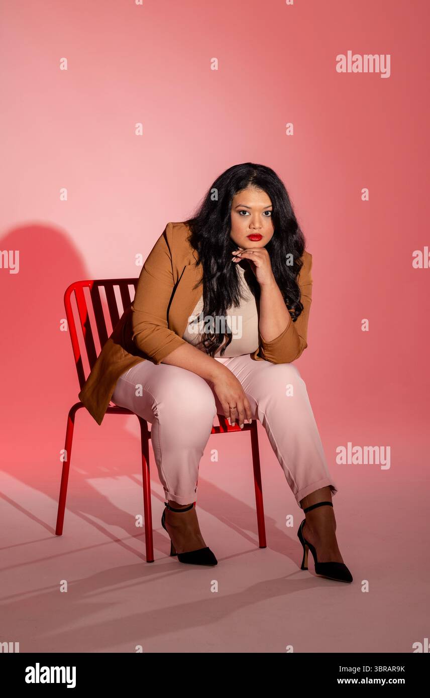 Donna seduta su una sedia in metallo rosso in studio con sfondo sfumato rosa e blazer marrone Foto Stock