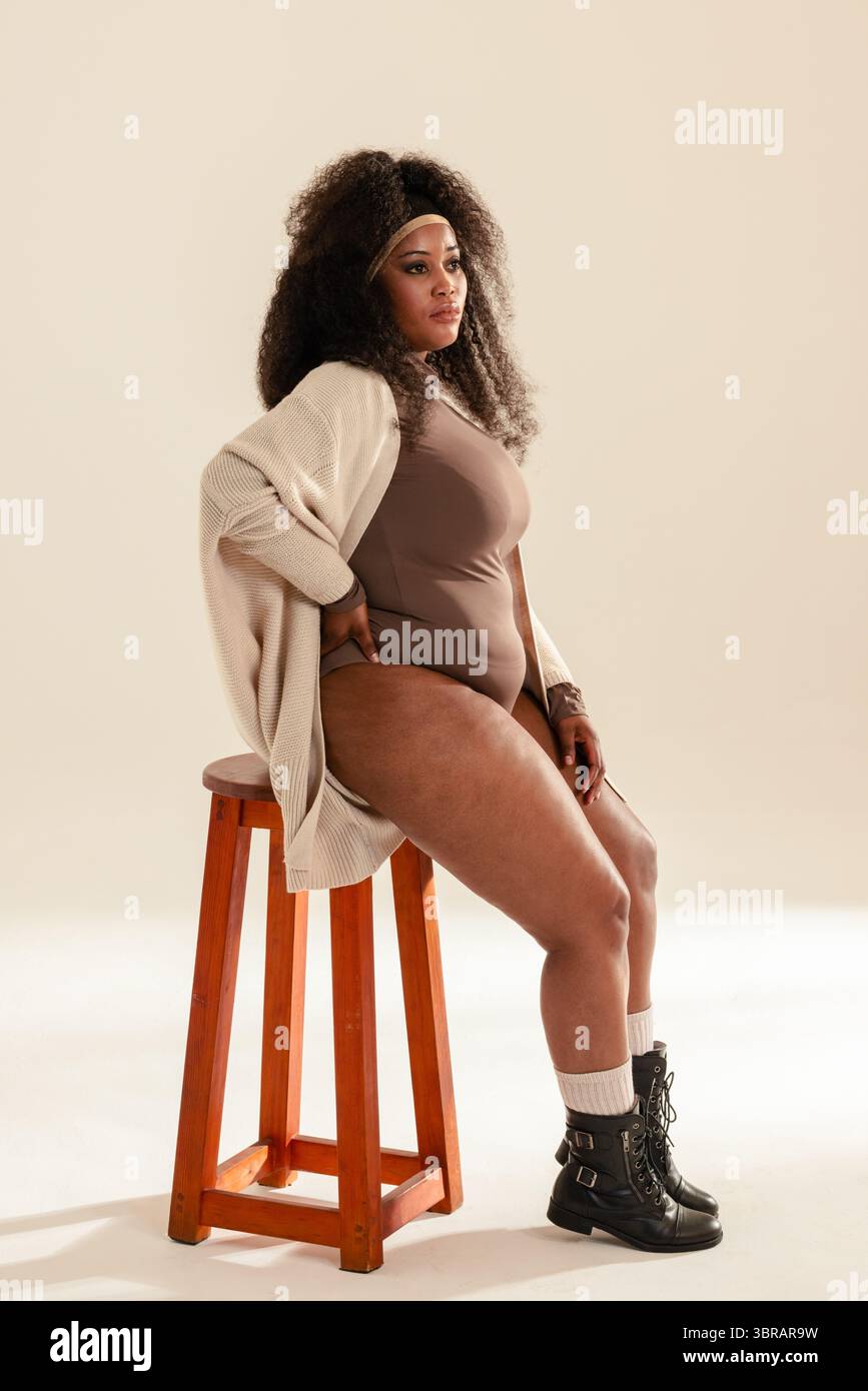 Donna afroamericana seduta in studio su uno sgabello che indossa un body e un cardigan con stivali da combattimento Foto Stock