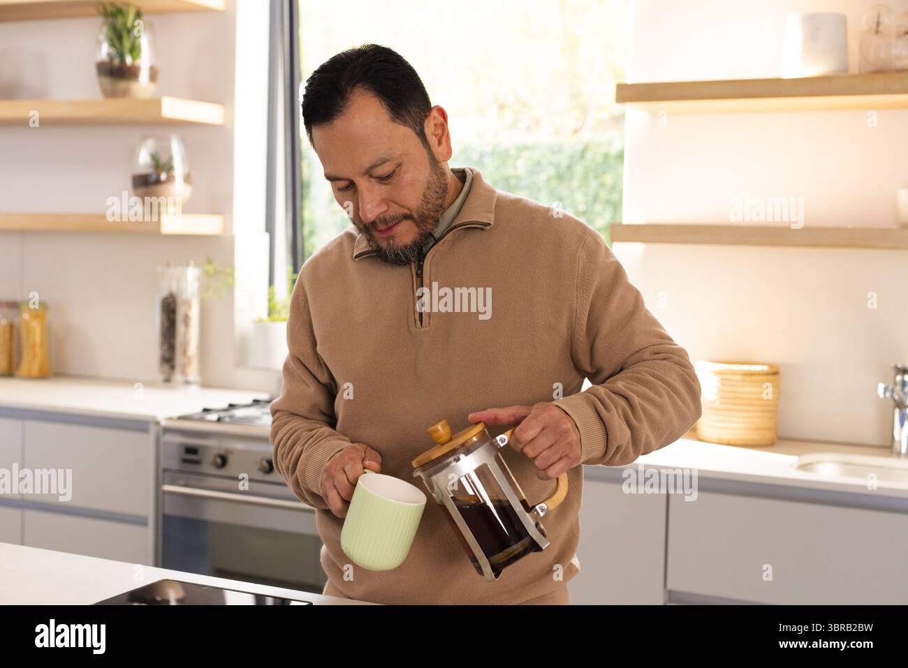 Uomo asiatico maturo che versa caffè dalla pressa francese in una tazza di ceramica verde nella moderna isola della cucina Foto Stock