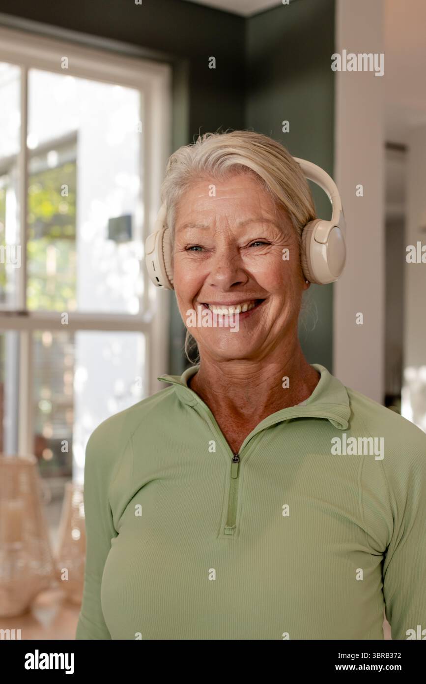 Donna anziana in salotto con cuffie wireless e zip verde sopra la finestra Foto Stock