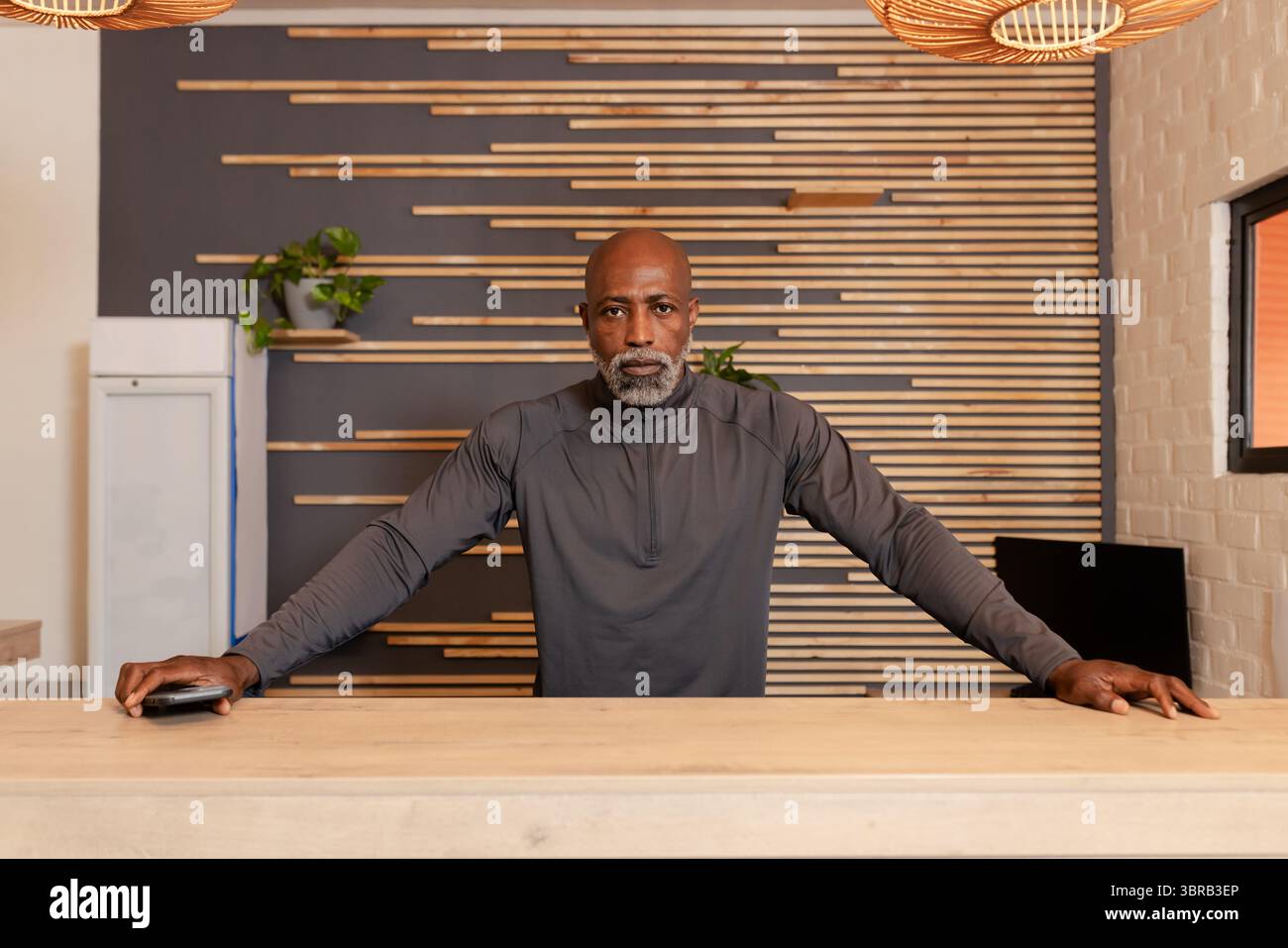 Uomo afro-americano in piedi dietro il banco ricevimento in legno chiaro nella lobby che tiene il dispositivo per carte di credito Foto Stock