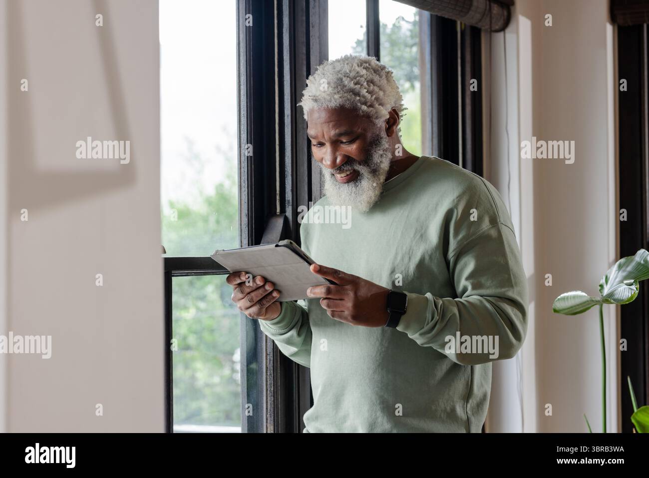Uomo afroamericano anziano che indossa uno smartwatch sorridente al tablet dalla finestra all'interno di casa, copia spazio Foto Stock