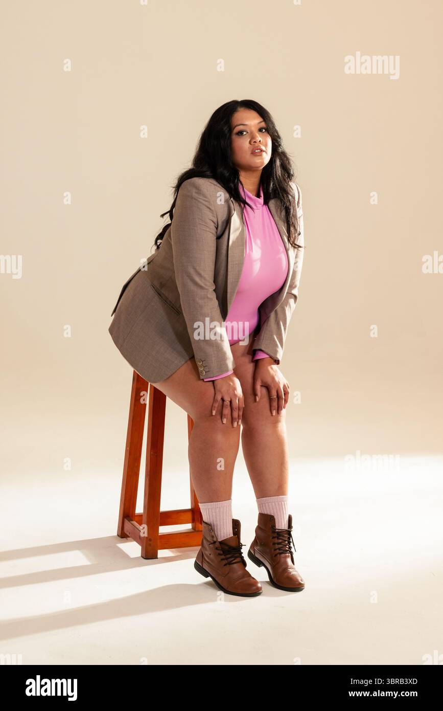 Donna di mezza età appoggiata su sgabello in legno in studio con stivali con top rosa blazer grigio Foto Stock