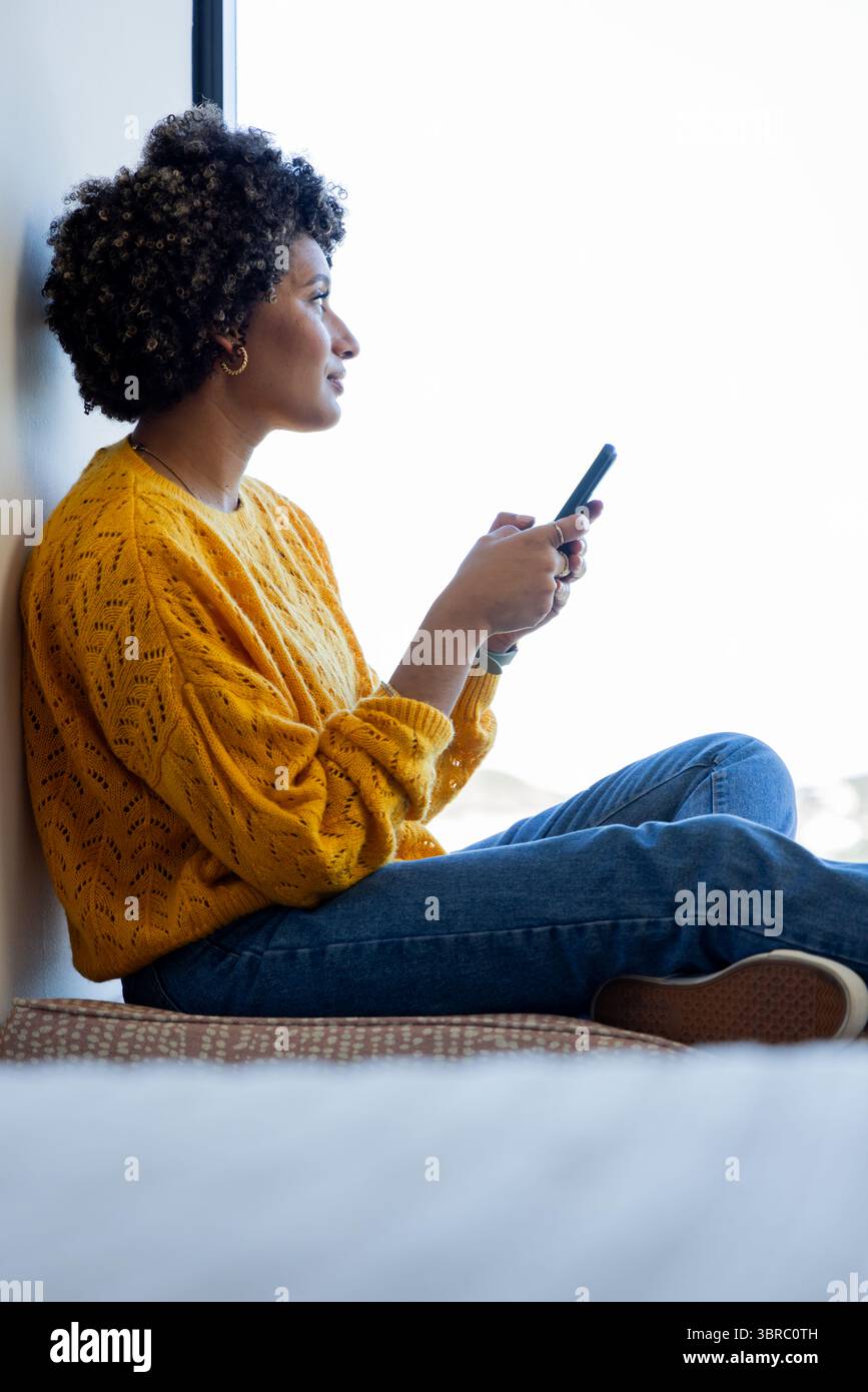 Donna afroamericana seduta su una tenera finestra con lo smartphone e guardando all'esterno, spazio per copiare Foto Stock
