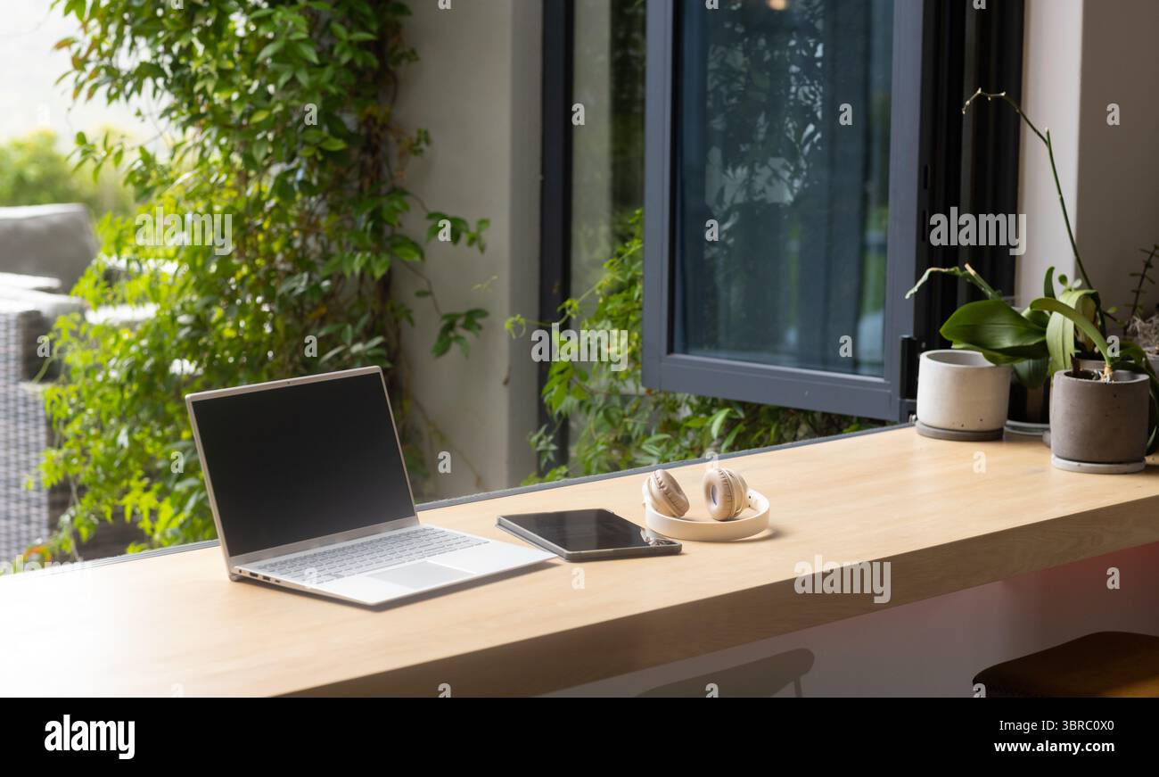 Computer portatile in argento aperto con tablet e cuffie su un bancone in legno vicino a una finestra aperta, con piante Foto Stock