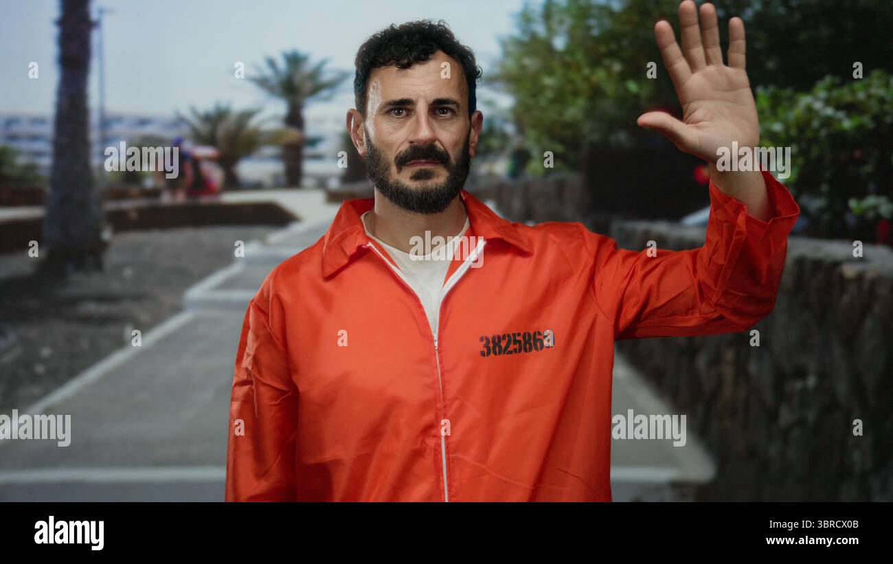Uomo ispanico nella tuta arancione della prigione all'aperto alzando la mano su una strada costeggiata da palme e vegetazione, creando un contrasto tra il confinamento A. Foto Stock