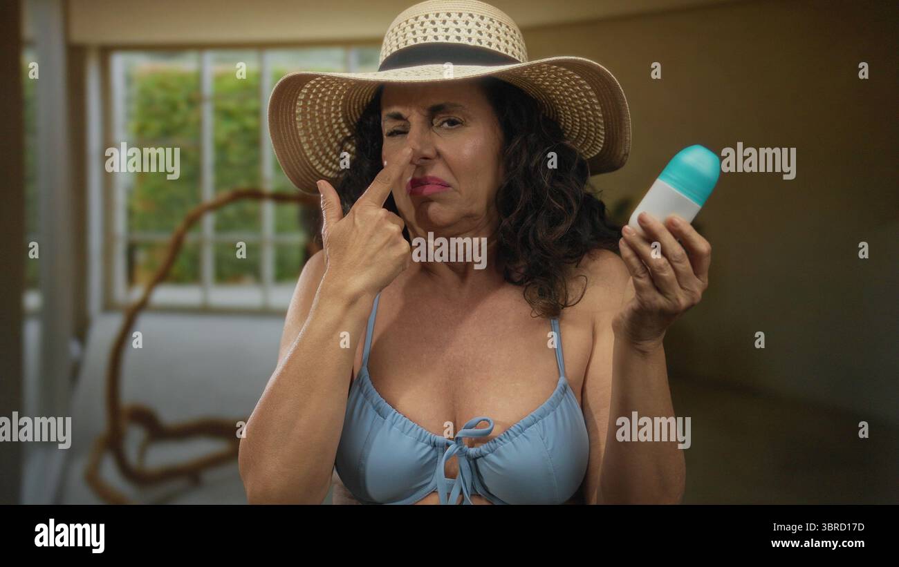Donna ispanica al chiuso che indossa bikini e cappello, che tiene il deodorante con un'espressione scontenta, mostrando una scena estiva all'interno di un ambiente interno. Foto Stock