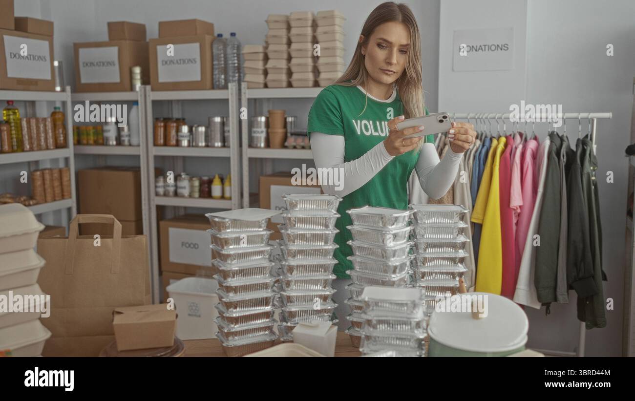 Donna volontaria che utilizza lo smartphone in un centro di beneficenza circondato da scatole per donazioni, contenitori per alimenti e scaffali per abbigliamento, sottolineando l'organizzazione dell'ambiente interno Foto Stock