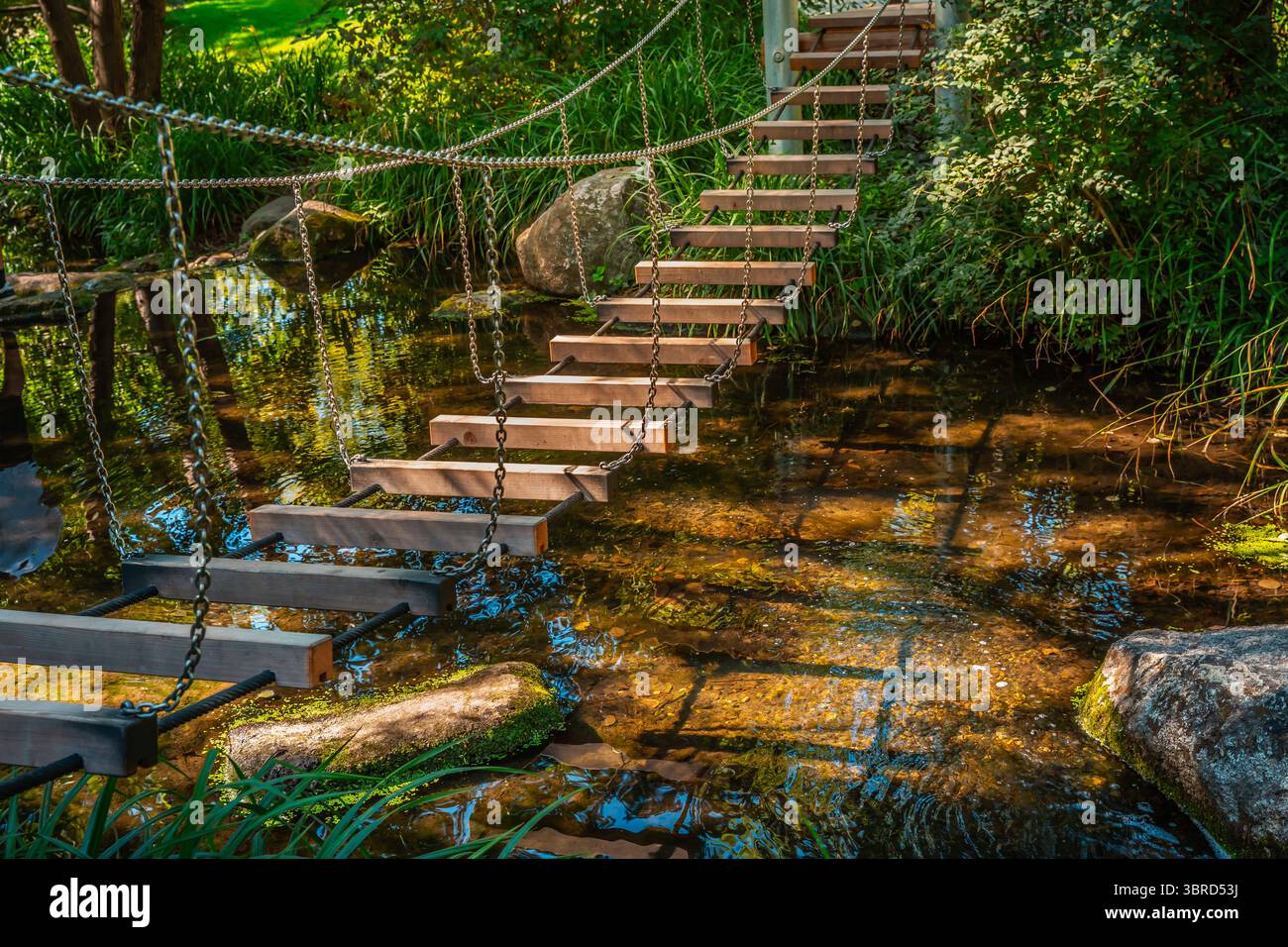 Piccolo ponte sospeso in un giardino o parco sul piccolo fiume o laghetto Foto Stock