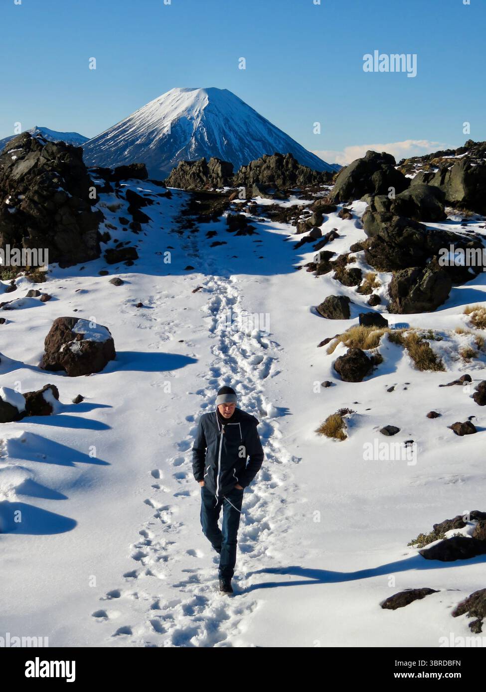 Un escursionista solitario cammina attraverso la neve fresca nel Parco Nazionale del Tongariro Leaves Footprints, con l'iconico cono del Monte Ngauruhoe che si erge alle spalle. Foto Stock