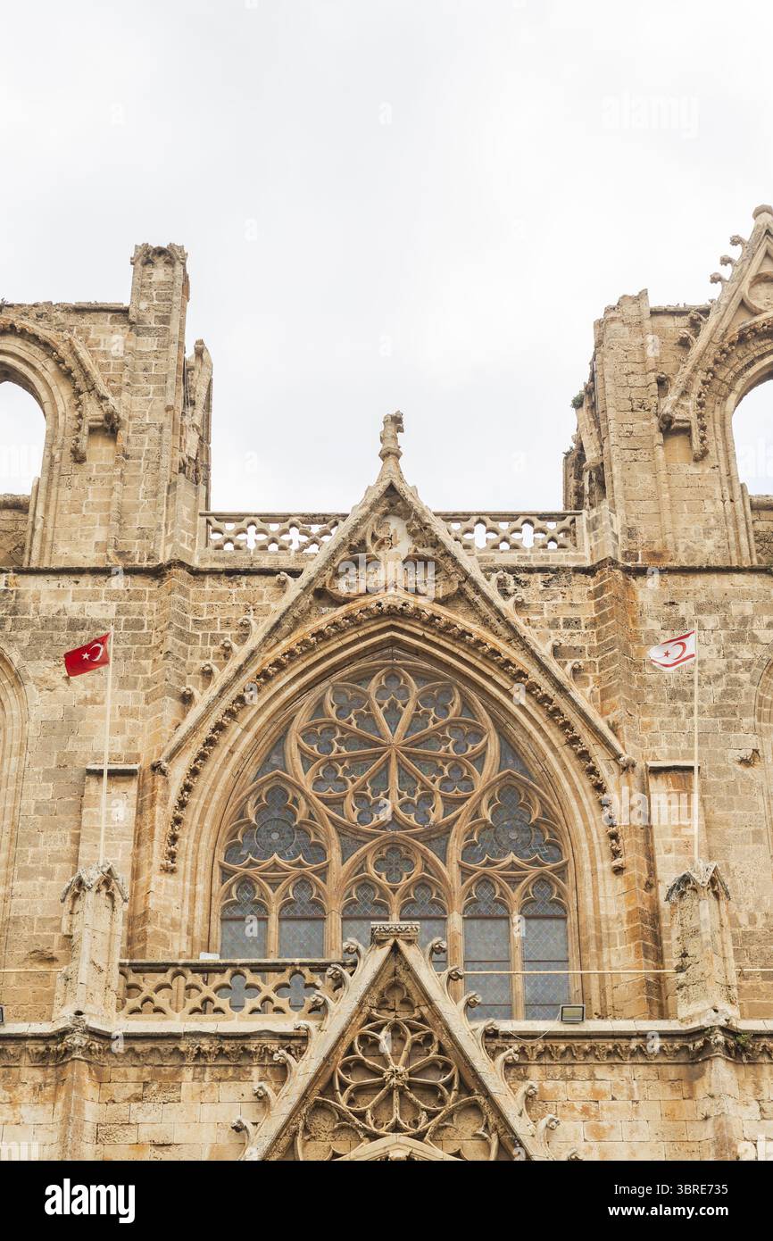 Vista della facciata sbriciolata di un edificio storico adornato con bandiere, le sue intricate pietre incise dal tempo sotto un cielo morbido, Varosha, Famagosta, Cipro. Foto Stock