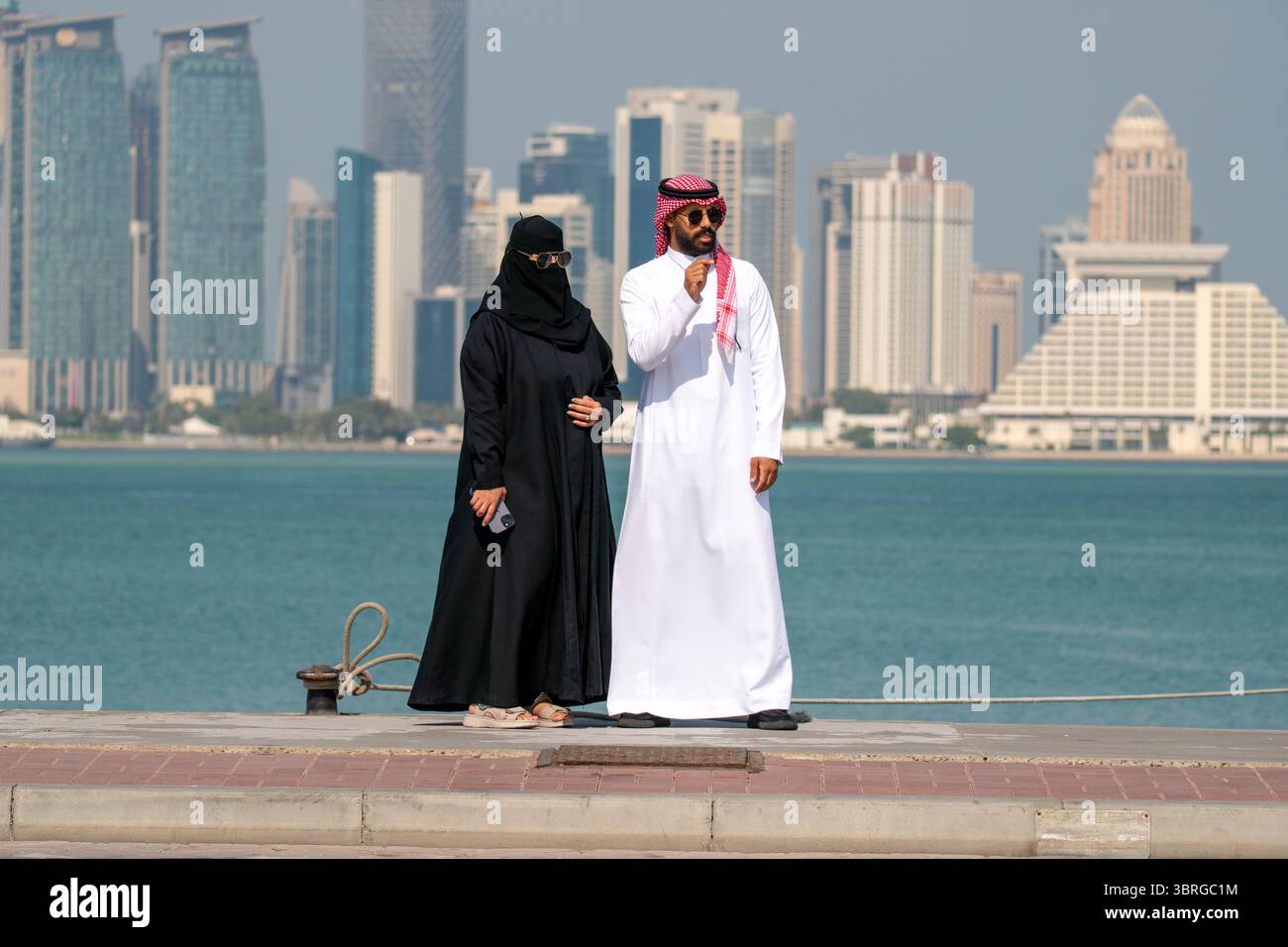 Una coppia si trova accanto all'acqua, godendosi il sole. Un uomo e una donna in abiti arabi tradizionali camminano lungo l'argine contro il dorso Foto Stock