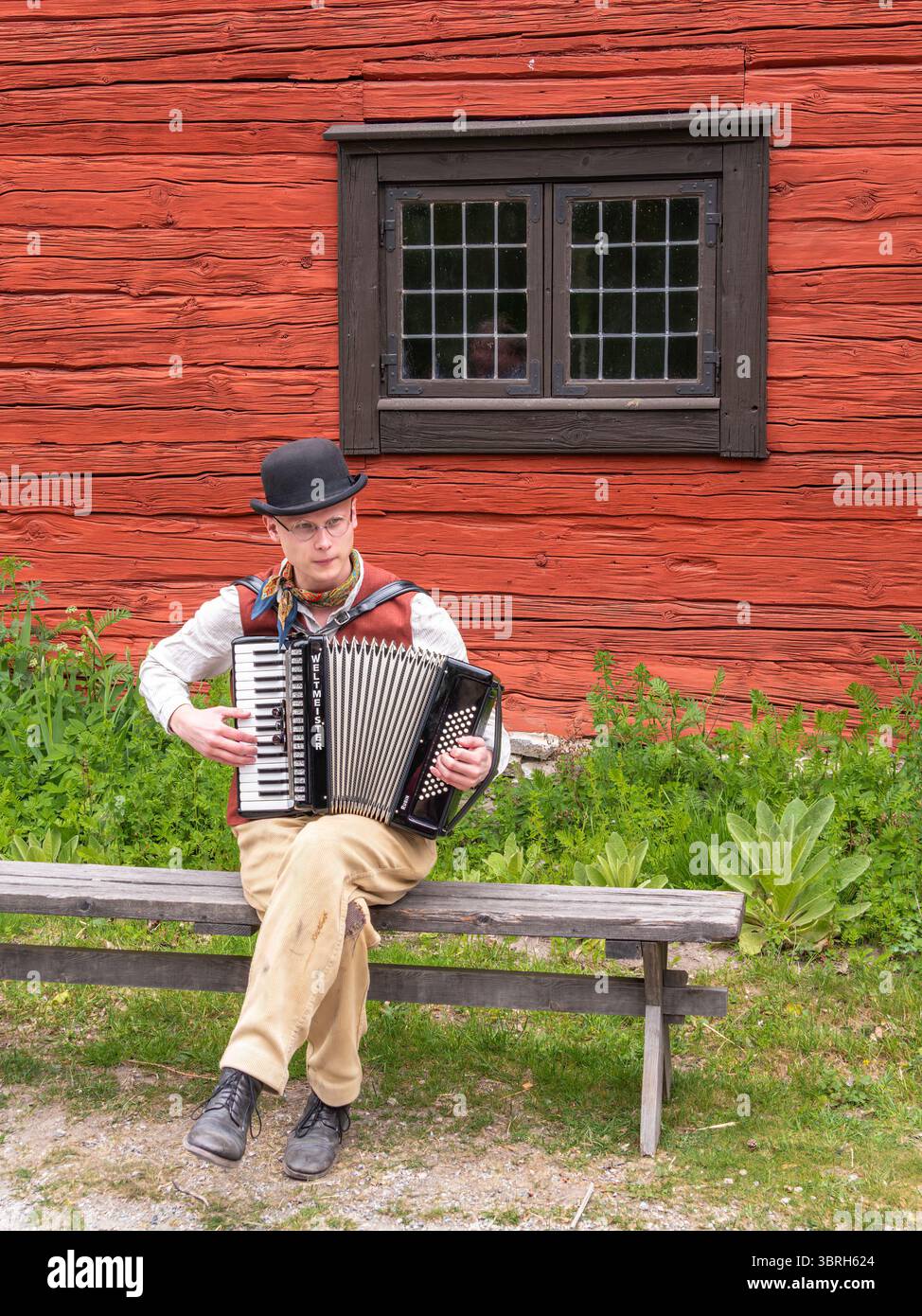 Sodertalje, Svezia - 6 giugno 2025: Un uomo che suona la fisarmonica seduto su una panchina di legno di fronte a un rustico edificio rosso. Riproduzione avvenuta in c Foto Stock